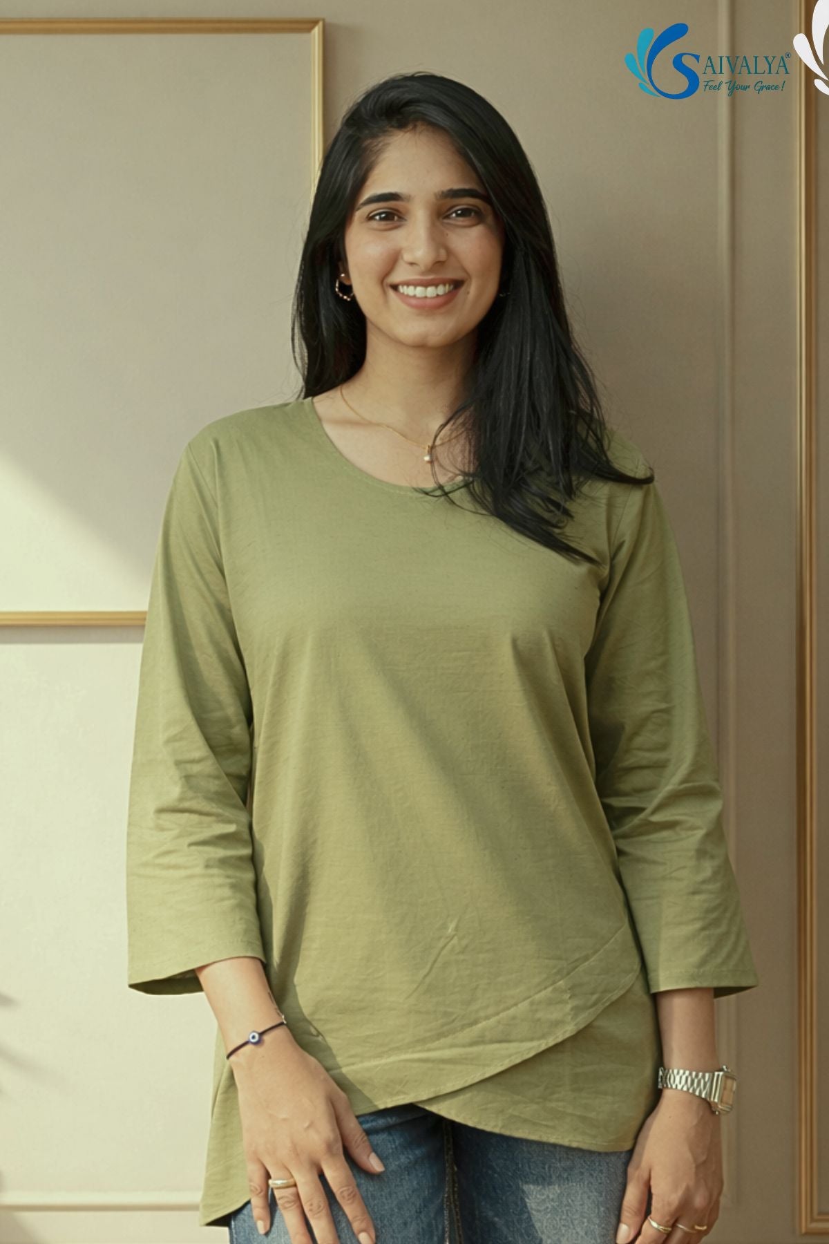 Medium front view of a smiling woman standing in a minimalist indoor studio setting with a gold frame in the background, wearing the Prakriti Sage Overlap Tunic featuring a light sage green color, a round neckline, three-quarter sleeves, and an asymmetric overlap tulip hemline, paired with blue denim jeans.