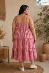 Back view of a smiling woman standing on a round jute rug in a warm bohemian room and looking over her shoulder, wearing the Raspberry Striped Tiered Midi Dress styled independently without the inner layer, highlighting its straight back neckline, thin straps, back smocking elastic details, and pink striped tiered flared skirt, paired with white sneakers.