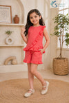 Front view of a smiling young girl posing with her hand in her pocket, wearing the coral pink Ruffle Charm Co-ord Set featuring a sleeveless asymmetric top with a diagonal ruffle overlay and matching ruffle-hem casual shorts.
