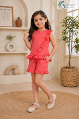 Front view of a smiling young girl posing with her hand in her pocket, wearing the coral pink Ruffle Charm Co-ord Set featuring a sleeveless asymmetric top with a diagonal ruffle overlay and matching ruffle-hem casual shorts.
