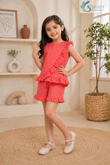 Three-quarter side profile view of a smiling young girl posing with her hand on her hip, wearing the coral pink Ruffle Charm Co-ord Set featuring a sleeveless asymmetric top with diagonal ruffle details and matching ruffle-hem casual shorts.