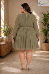 Full-length back view of a plus-size woman looking over her shoulder in a warm, elegantly styled indoor setting with a wooden console table and potted palm tree in the background, wearing the Sage Georgette Pleated Muse Dress highlighting its light olive-sage green plissé pleated fabric, high mock neckline with a back keyhole button closure, long sheer sleeves with ruffled cuffs, matching fabric waist tie, and a flared pleated skirt, paired with nude pointed heels and a beige clutch.