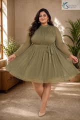 Full-length front view of a smiling plus-size woman holding out the sides of her dress to showcase a wide flare in a warm, elegantly styled indoor setting with a wooden console table and potted palm tree in the background, wearing the Sage Georgette Pleated Muse Dress featuring a light olive-sage green plissé pleated fabric, a high mock neckline, long sheer sleeves with ruffled cuffs, a matching fabric waist tie, and an expansive flared pleated skirt, paired with nude pointed heels.