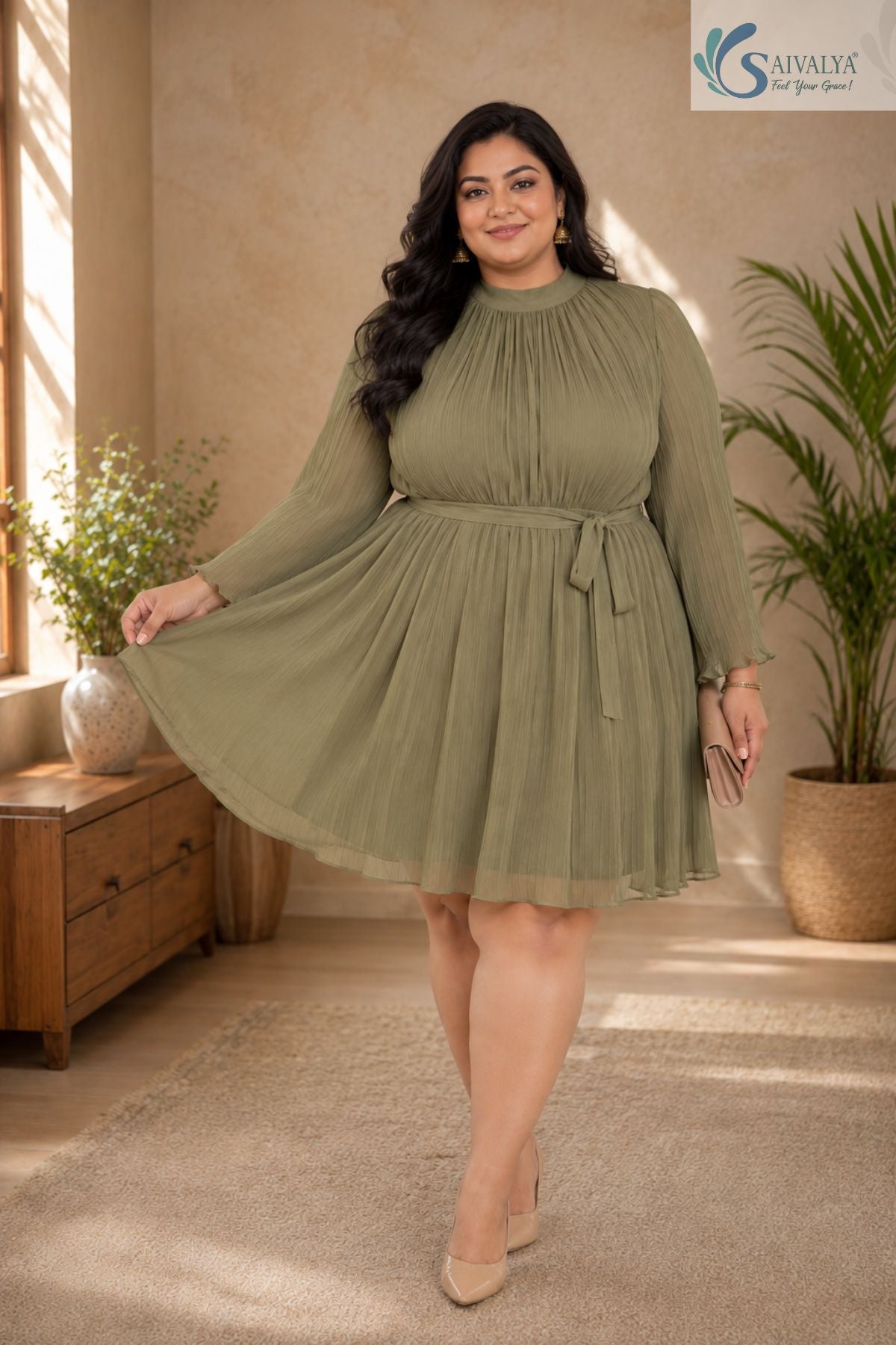 Full-length front view of a smiling plus-size woman posing gracefully in a warm, elegantly styled indoor setting with a wooden console table and potted palm tree in the background, wearing the Sage Georgette Pleated Muse Dress featuring a light olive-sage green plissé pleated fabric, a high mock neckline, long sheer sleeves with ruffled cuffs, a matching fabric waist tie, and a flared pleated skirt, paired with nude pointed heels and a beige clutch.