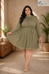 Full-length front view of a smiling plus-size woman posing gracefully in a warm, elegantly styled indoor setting with a wooden console table and potted palm tree in the background, wearing the Sage Georgette Pleated Muse Dress featuring a light olive-sage green plissé pleated fabric, a high mock neckline, long sheer sleeves with ruffled cuffs, a matching fabric waist tie, and a flared pleated skirt, paired with nude pointed heels and a beige clutch.