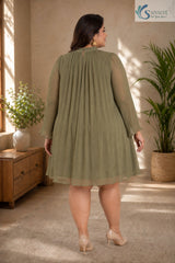Full-length back view of a plus-size woman looking over her shoulder in a warm, elegantly styled indoor setting with a wooden console table and potted palm tree in the background, wearing the Sage Georgette Pleated Muse Dress styled without its belt to showcase a loose, flowing babydoll silhouette in light olive-sage green plissé pleated fabric with a high mock neckline featuring a back keyhole button closure and long sheer sleeves with ruffled cuffs, paired with nude pointed heels.