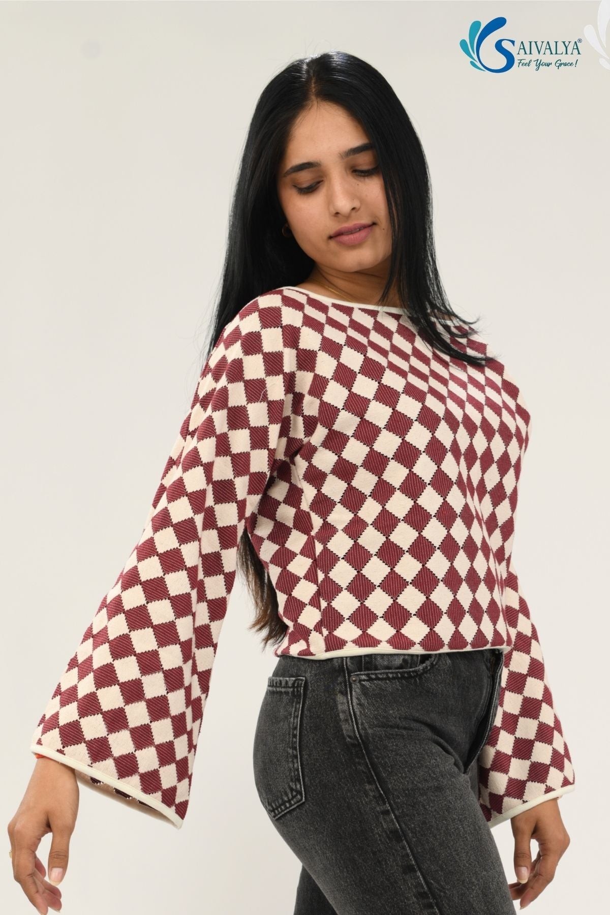 A woman wearing the Shatranj Maroon Bell Top, featuring a maroon and cream checkered harlequin pattern with long bell sleeves, paired with dark grey denim jeans against a plain white background.