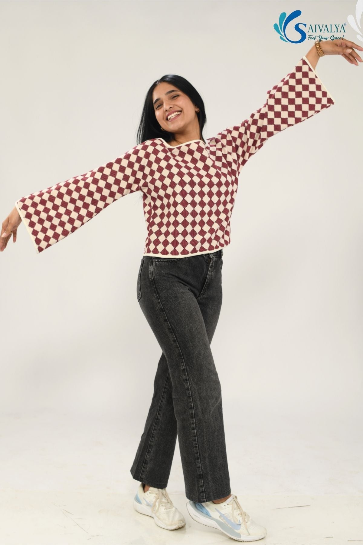 Full-length view of a woman smiling and posing with arms outstretched to show the dramatic flare of the Shatranj Maroon Bell Top. She is wearing the maroon and cream checkered top with high-waisted dark grey wide-leg jeans and white sneakers against a bright studio background.