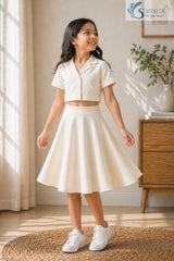 Full-length view of a smiling young girl looking to the side, wearing a cream Slub Cotton Co-ord set featuring a short-sleeve button-up crop shirt and a flared midi skirt with white sneakers.