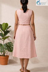 Full-length back view of a woman standing facing away from the camera in a bright, minimalist indoor studio setting with a potted fiddle-leaf fig plant in the background, wearing The Scalloped Blush Midi Set highlighting its sleeveless powder pink cotton crop top with a scalloped petal-cut hemline, a central vertical back seam, and a matching high-waisted, A-line midi skirt with subtle back pleating details, completed with tan leather flat slides.