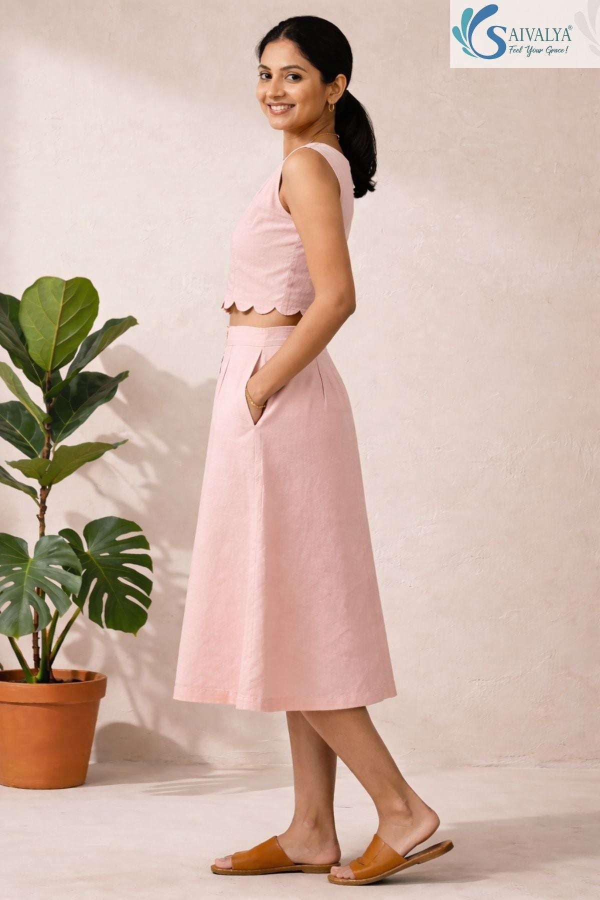 Full-length side view of a smiling woman posing with a hand in her pocket in a bright, minimalist indoor studio setting with a potted fiddle-leaf fig plant in the background, wearing The Scalloped Blush Midi Set featuring a sleeveless powder pink cotton crop top with a delicate scalloped hemline, paired with a matching high-waisted, A-line midi skirt with side pockets, completed with tan leather flat slides.
